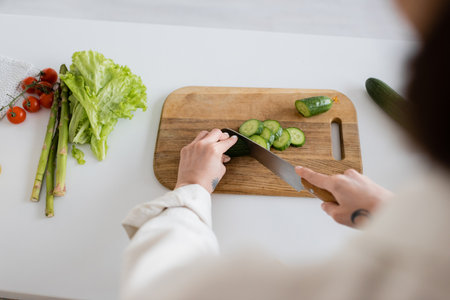 Top view of tattooed woman cutting cucumber near vegetables in kitchenの写真素材