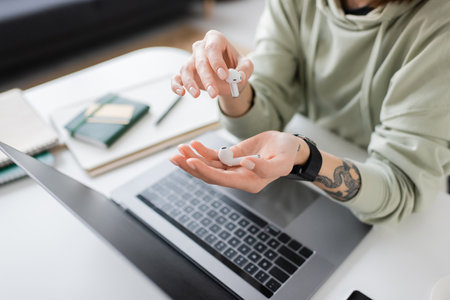 Cropped view of tattooed freelancer holding earphones near laptop with blank screen on table at homeの写真素材