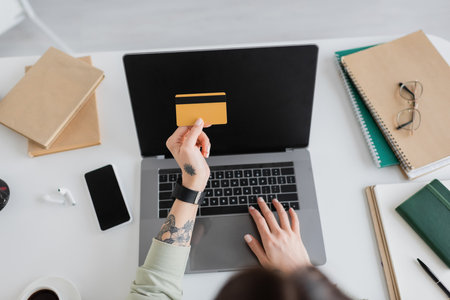 Cropped view of tattooed woman holding credit card near gadgets and notebooks at home