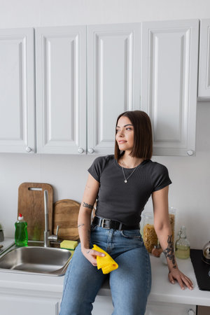 Smiling brunette woman holding rag while sitting on worktop in kitchenの写真素材