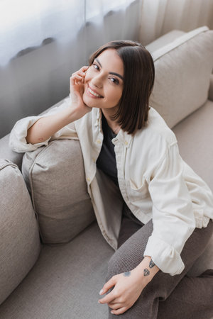Overhead view of smiling tattooed woman looking at camera while sitting on couch at home