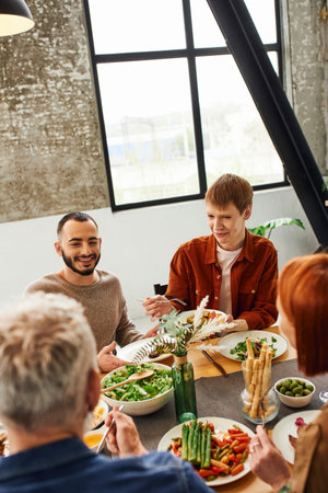 redhead gay man smiling near bearded boyfriend and blurred parents with delicious meal served in...