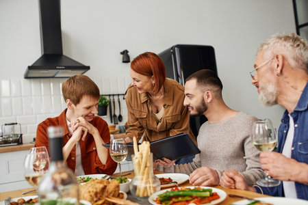 cheerful redhead woman showing family photo album to smiling son with gay boyfriend near delicious supper served in kitchenの写真素材
