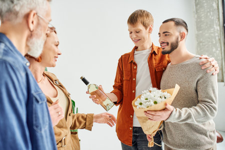 cheerful gay couple holding flowers and wine bottle near parents at homeの写真素材