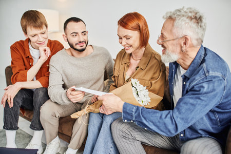 smiling gay man giving wedding invitation to parents near boyfriend on sofa in living roomの写真素材