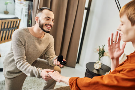 cheerful bearded man holding jewelry box near blurred boyfriend while making marriage proposal at homeの写真素材