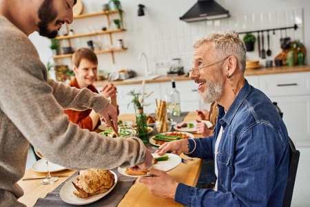 smiling bearded man looking at gay man serving chicken during supper with family in kitchenの写真素材
