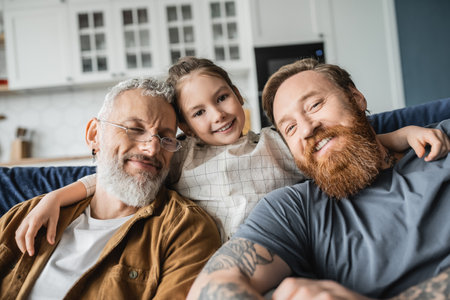 Smiling kid hugging bearded homosexual fathers on couch at home