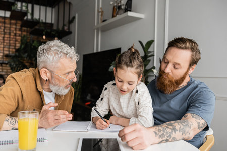 Preteen child writing on notebook near homosexual dads and digital tablet at homeの写真素材
