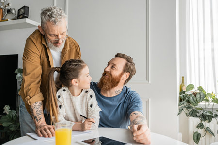 Positive gay man looking at partner near daughter doing homework and digital tablet at homeの写真素材
