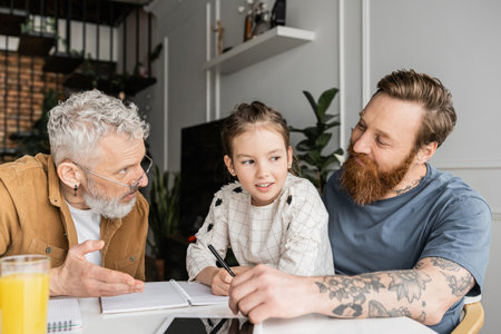 Gay fathers talking to smiling preteen daughter doing homework at homeの写真素材