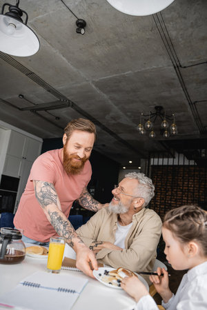 Cheerful gay man putting plate with pancakes on table near daughter and partner at home