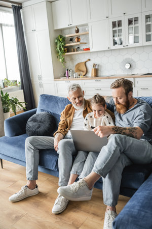 Homosexual fathers using laptop with cheerful preteen daughter at home