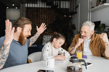Excited gay dads sitting near preteen daughter writing on notebook at homeの写真素材