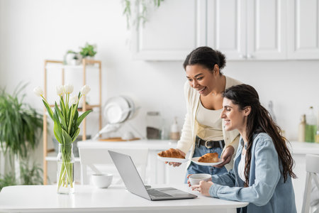 happy multiracial woman holding plates with croissants near girlfriend working on laptop from homeの写真素材