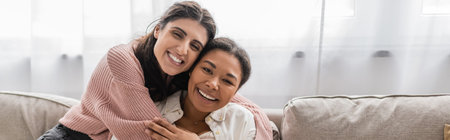 happy lesbian woman hugging multiracial partner and sitting on couch, banner