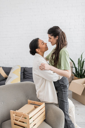 positive lesbian woman hugging happy multiracial girlfriend near wooden box in living room
