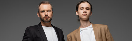 well dressed teenager boy and father in suits looking at camera isolated on grey, banner