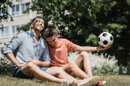 teenage boy sitting near joyful father and holding soccer ball in green summer parkの写真素材