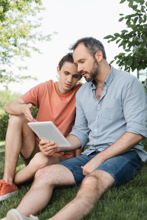 bearded dad and teenager son looking at digital tablet while sitting in green parkの写真素材
