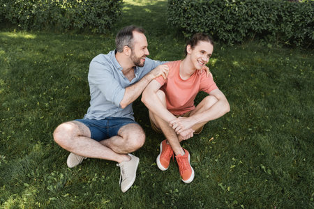 overhead view of happy bearded father hugging teenage son while sitting together on green lawnの写真素材