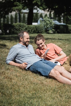 bearded man smiling while using smartphone and resting on lawn near teenage son after playing...