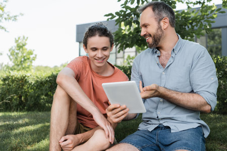 bearded dad and happy teenager son looking at digital tablet while sitting in green parkの写真素材
