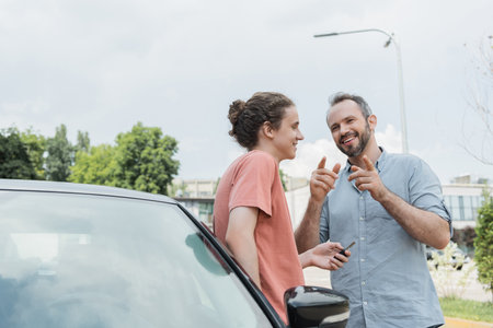 happy bearded dad pointing with fingers near teenager son standing near car with keysの写真素材