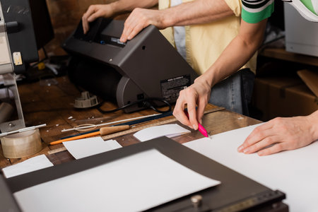 cropped view of typographer cutting paper with knife near colleague in print center