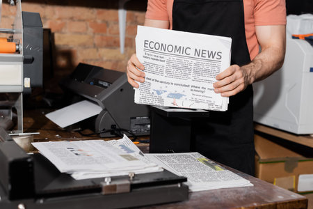 cropped view of young typographer in apron holding newspapers with economic news