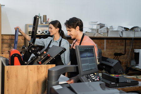 cheerful man and woman in aprons using professional print plotter while working together in print...