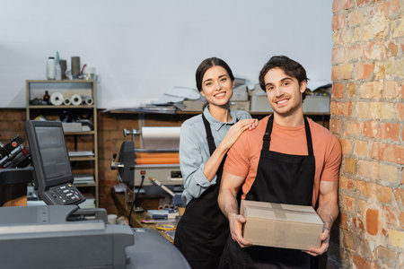 cheerful typographer leaning on happy colleague with carton box
