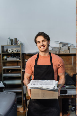 cheerful man in typographer holding carton box with newspapers and smiling in print center