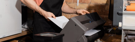 cropped view of worker in apron holding paper near printer and fresh newspapers in print center,...