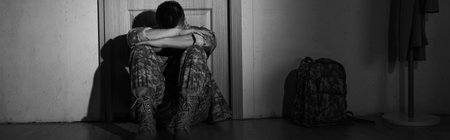 Black and white photo of depressed soldier sitting near backpack and door in hallway at home, bannerの写真素材