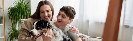 Cheerful and young gay couple in casual clothes petting and looking at furry Australian shepherd dog...