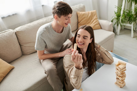 High angle view of cheerful young gay couple in casual clothes playing wood blocks game on table while spending time together in living room at homeの写真素材