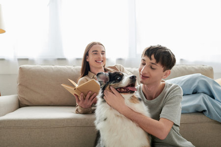 happy young gay man with long hair holding book and resting on comfortable sofa next to his boyfriend with tattoo cuddling Australian shepherd dog in modern apartmentの写真素材