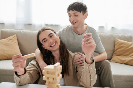 Smiling gay man hugging and looking at long haired and tattooed boyfriend while playing blurred wood blocks game on table near couch in living room at homeの写真素材
