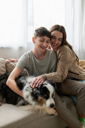 Long haired and tattooed gay man petting furry Australian shepherd dog near smiling partner in...