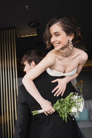 groom in black formal wear lifting joyful bride in white wedding dress and luxurious jewelry holding bridal bouquet with flowers while standing in hotel lobbyの写真素材
