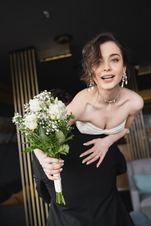 groom in black formal wear lifting pretty bride in white wedding dress and luxurious jewelry holding bridal bouquet with flowers while standing in hotel lobbyの写真素材