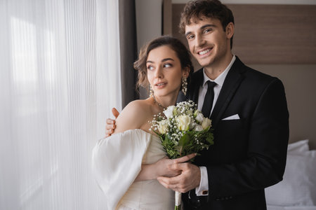 cheerful groom in classic formal wear hugging elegant bride in jewelry, white dress with bridal bouquet while standing together in modern hotel room after wedding ceremonyの写真素材