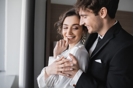 groom in classic black suit embracing happy young bride in jewelry and white dress while standing together in modern hotel room during their honeymoon on wedding day, joyful newlywedsの写真素材