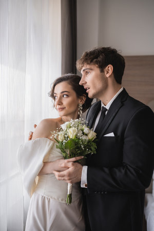amazed groom in classic formal wear standing with opened mouth next to elegant young bride in jewelry, white dress with bridal bouquet in modern hotel room after wedding ceremonyの写真素材