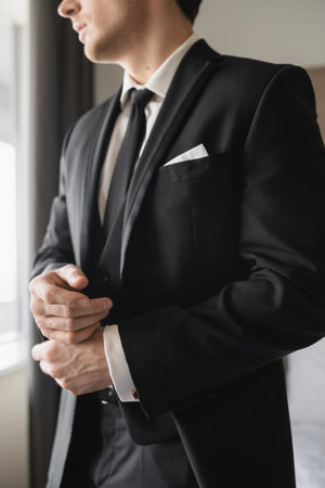 cropped view of groom in formal wear with classy black tie and white shirt standing in modern hotel room while adjusting handcuffs, groom on wedding day, special occasionの写真素材
