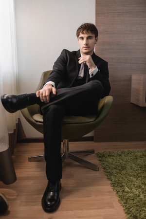 young man in black formal wear with tie and classic shoes sitting on comfortable armchair and looking at camera on wedding day, posing in modern hotel room, good looking groomの写真素材