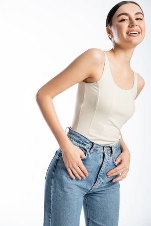 appealing and young woman with natural makeup, brunette hair and perfect skin smiling while posing in tank top and denim jeans on white backgroundの写真素材