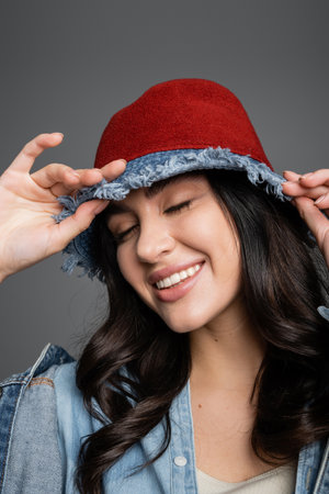 portrait of happy woman with closed eyes and flawless natural makeup posing in panama hat and denim jacket and smiling on grey backgroundの写真素材