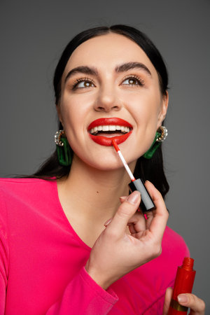 alluring woman with shiny brunette hair and trendy earrings applying red lip gloss and smiling while posing in magenta dress isolated on grey backgroundの写真素材